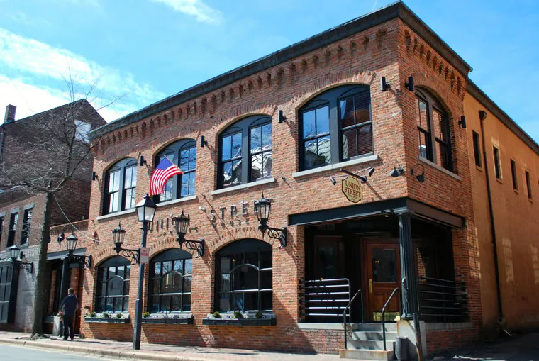 A two-story red brick building with large arched windows, black shutters, and an American flag. Three black lanterns hang above the entrance, and a person walks by on the sidewalk under a blue sky.