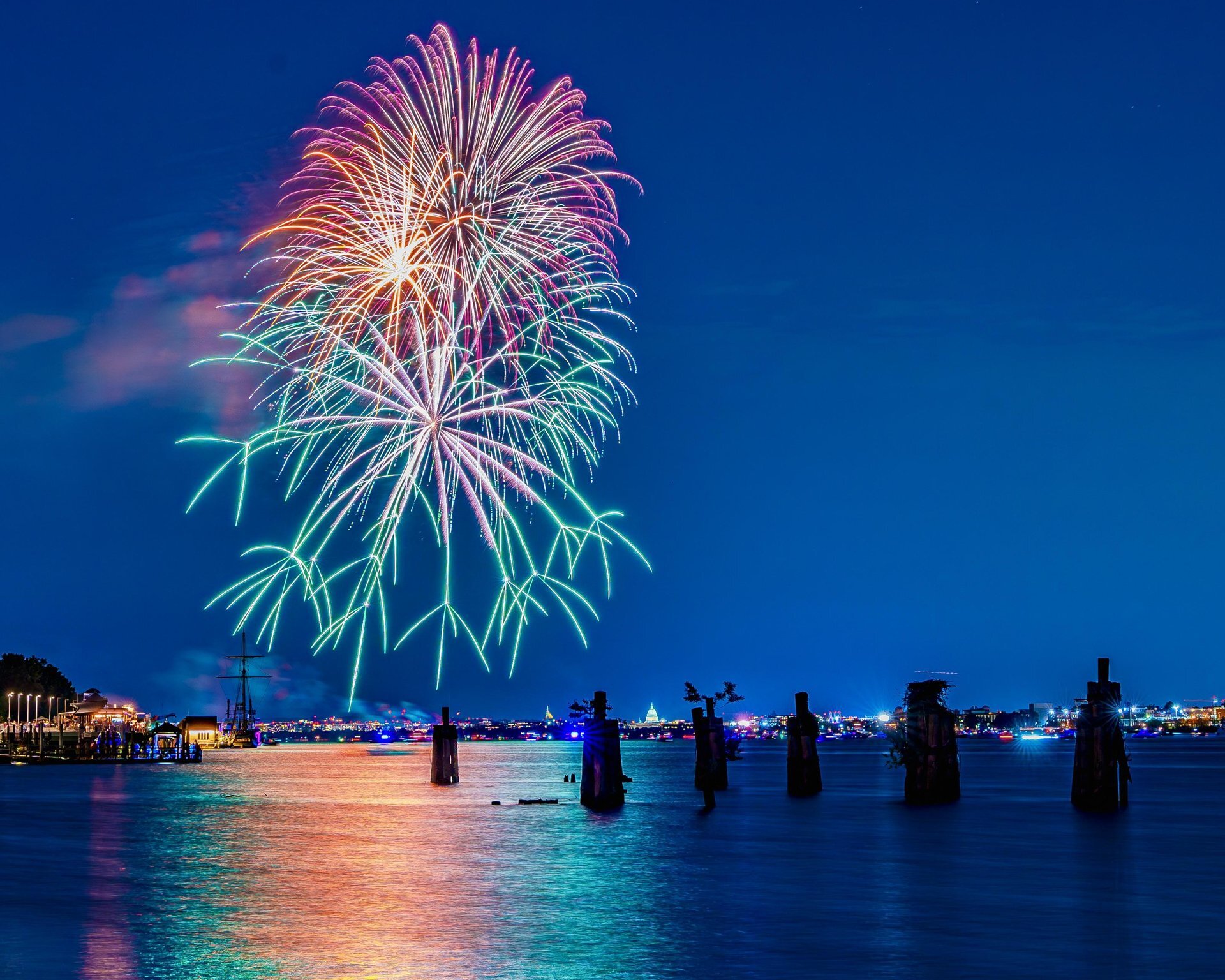 Colorful fireworks burst over a calm waterfront at night, reflecting on the water. Silhouetted posts and distant city lights are visible under a clear, deep blue sky.