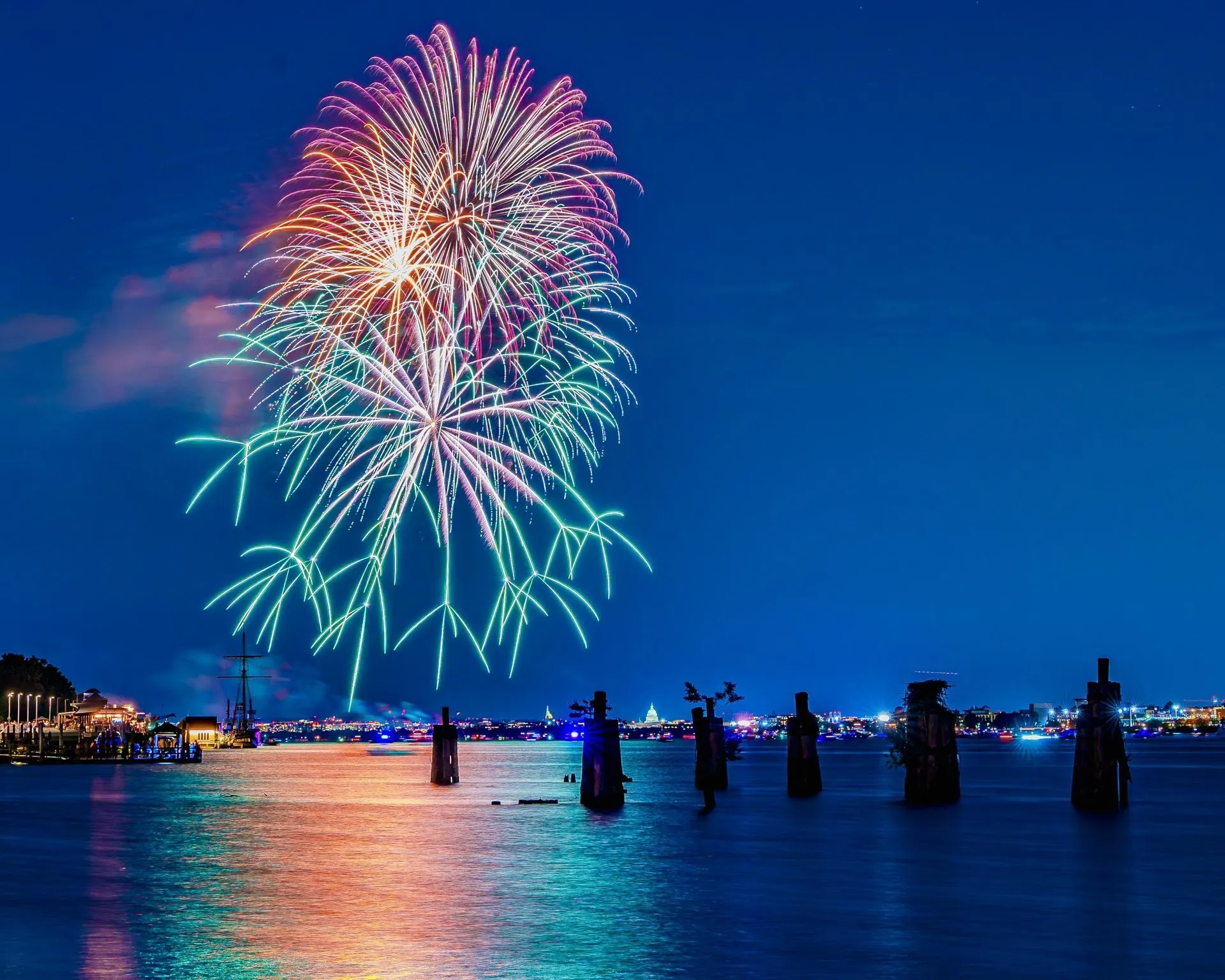 Colorful fireworks burst over a calm waterfront at night, reflecting on the water. Silhouetted posts and distant city lights are visible under a clear, deep blue sky.