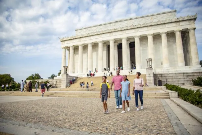 A family of four walks in front of the Lincoln Memorial on a sunny day, with other visitors on the steps and around the monument. The sky is partly cloudy.