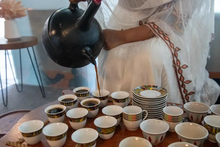 A person in a white traditional outfit pours coffee from a black pot into small, colorful cups arranged on a wooden table, preparing an Ethiopian coffee ceremony.