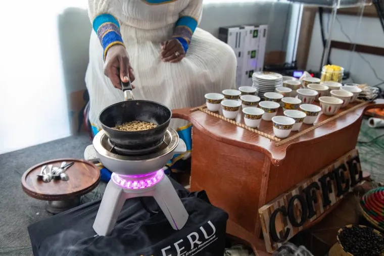 A woman in traditional attire roasts coffee beans in a black pan over an open flame. Nearby, a wooden tray holds many small cups, likely for a coffee ceremony. The word “COFFEE” is displayed on the tray.