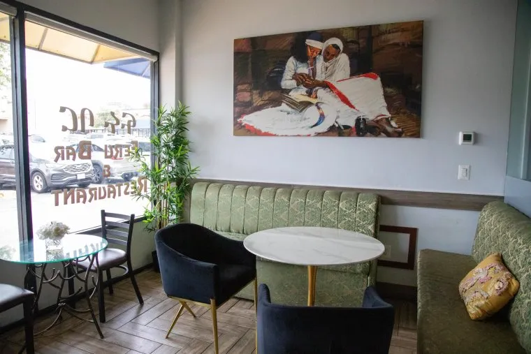 A cozy restaurant seating area with green upholstered benches, black chairs, round white tables, and a large painting of two people in traditional attire on the wall. Sunlight comes through a window with lettering.