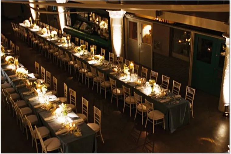 A large banquet hall set for an event, with long tables arranged in a U-shape, covered with tablecloths, white chairs, floral centerpieces, and glowing candlelight. The room is warmly lit and elegantly decorated.