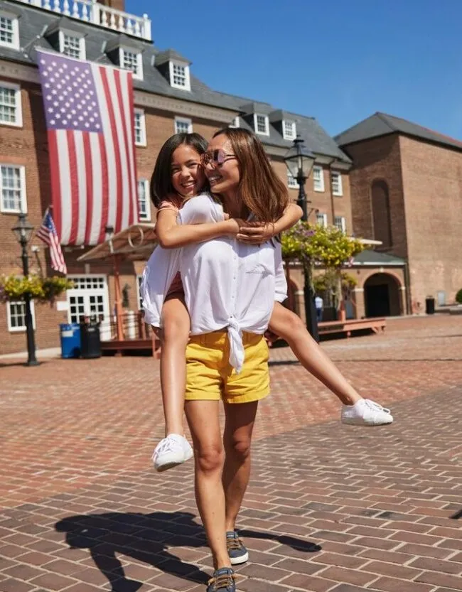 Two women smiling, one giving the other a piggyback ride in a sunny brick courtyard, with large American flags and a historic brick building in the background.