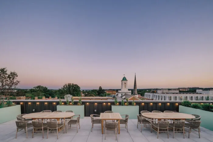 Rooftop patio with modern tables and chairs, overlooking a townscape with trees and historic buildings at sunset. A church steeple and clock tower are visible against a clear evening sky.