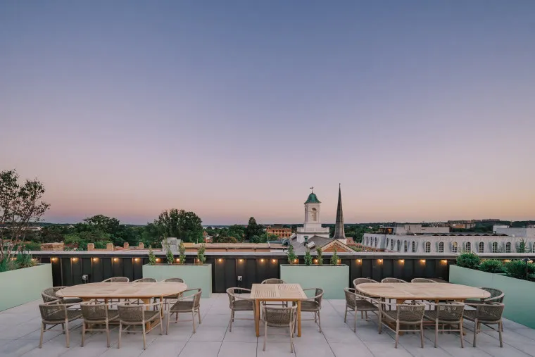 Rooftop patio with modern tables and chairs, overlooking a townscape with trees and historic buildings at sunset. A church steeple and clock tower are visible against a clear evening sky.