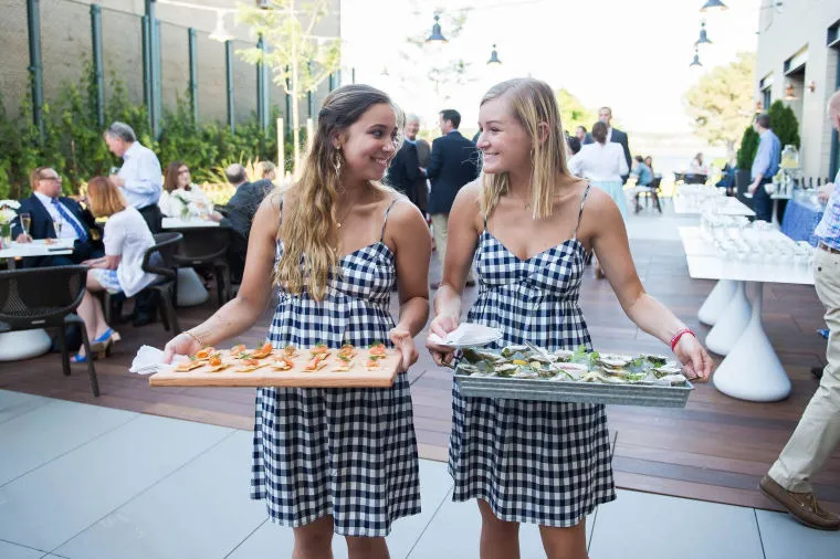 Two women in matching checkered dresses carry trays of appetizers at an outdoor event. People are seated at tables in the background, and the setting appears to be a terrace or patio with modern decor.