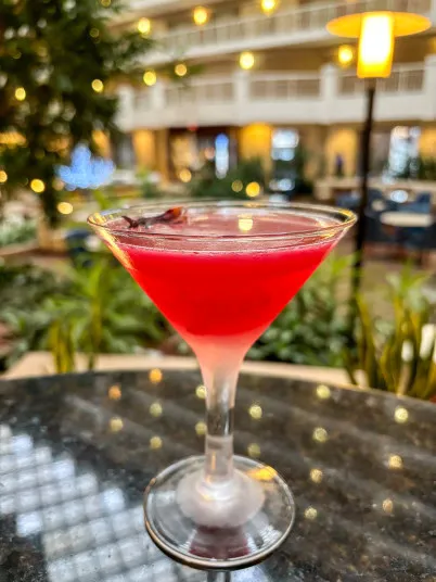 A vibrant red cocktail in a martini glass sits on a round, dark tabletop with a blurred background of greenery, warm string lights, and indoor seating in an atrium setting.