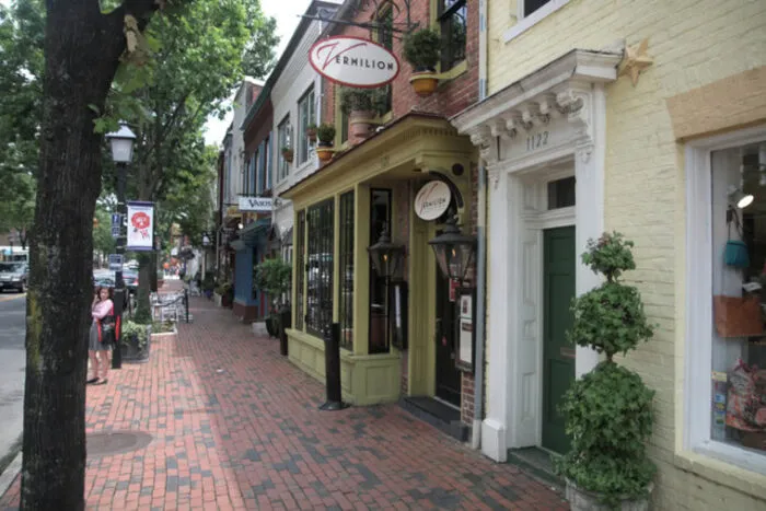 A brick sidewalk lined with trees and historic storefronts, including a shop with a green door and a sign reading Vermilion, on a quaint, lively street. People are walking and sitting at outdoor tables.