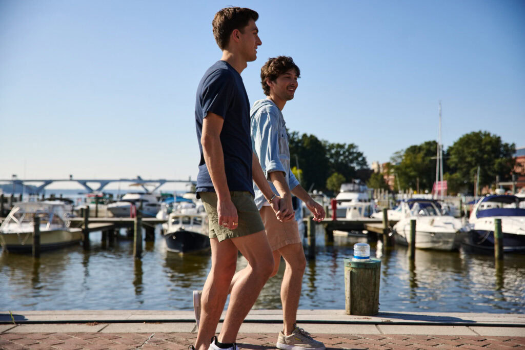 Two young men are walking along a marina on a sunny day, with boats docked in the water behind them and clear blue sky overhead.