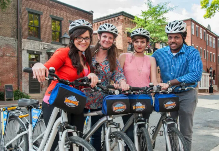 Four people wearing helmets stand with bicycles on a city street, smiling at the camera. The bikes have blue bags labeled “Bike and Roll.” Brick buildings and trees are visible in the background.