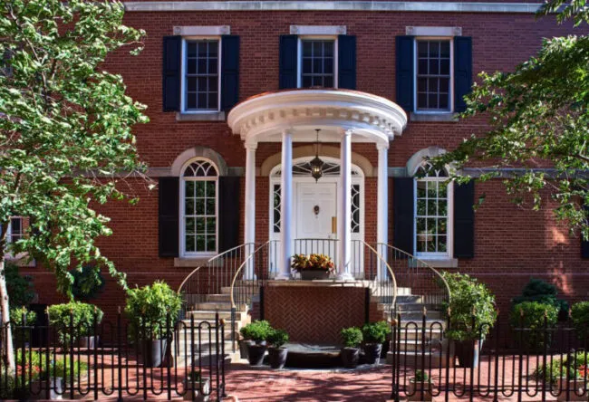 A brick house with black shutters, arched windows, and a white columned porch with a round roof and steps leading to a white front door, surrounded by trees and potted plants.