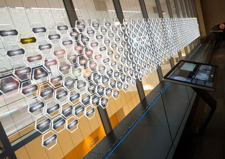 A long, illuminated wall display featuring interconnected hexagonal panels with text, resembling a digital network or flowchart, is shown in a modern indoor space with glass walls and information kiosks.