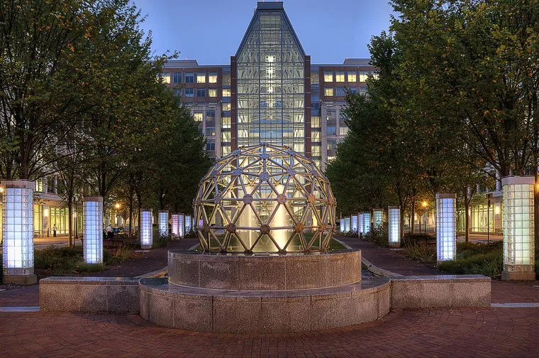 A large glass dome sculpture sits in a fountain, surrounded by trees and illuminated columns, in front of a modern office building with a tall glass atrium at dusk.