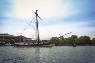 A historic tall ship with sails furled is docked on a calm river, surrounded by trees and buildings under a partly cloudy sky.