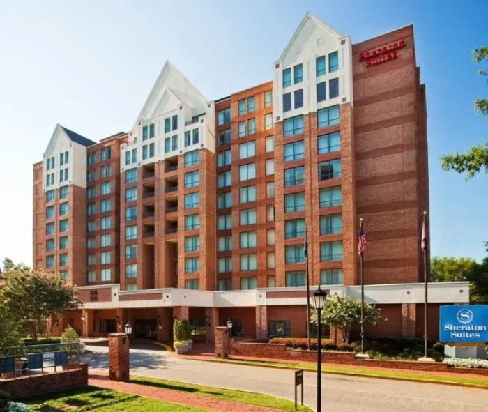 A multi-story red brick hotel building with white gabled rooftops, a Sheraton Suites sign at the front, and American flags near the entrance on a sunny day.
