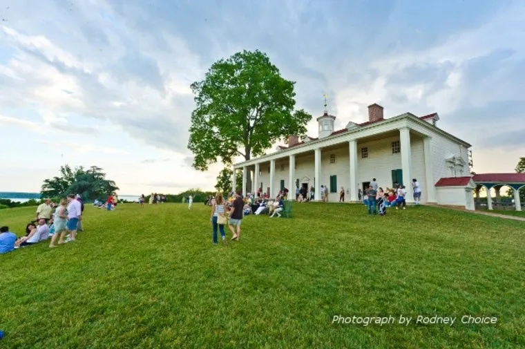 A large group of people relax and gather on the grassy lawn in front of a historic white mansion with columns and a cupola, under a partly cloudy sky. Text in the corner credits the photo to Rodney Choice.
