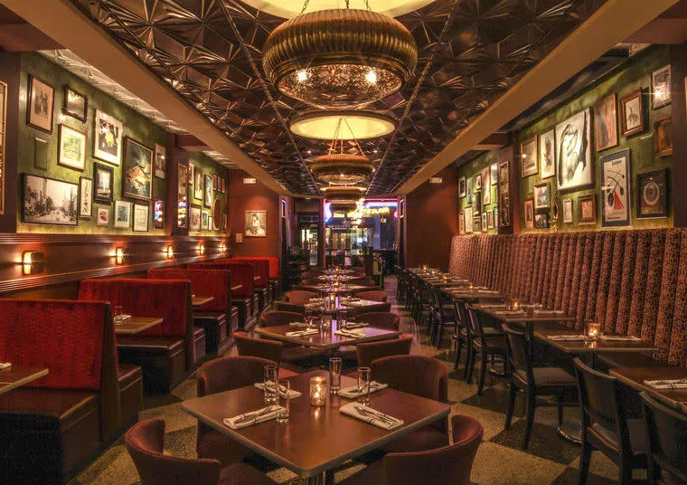 Cozy restaurant interior with dim lighting, red upholstered booths on the left, patterned banquettes on the right, neatly set tables in between, framed art on green walls, and ornate pendant lights overhead.