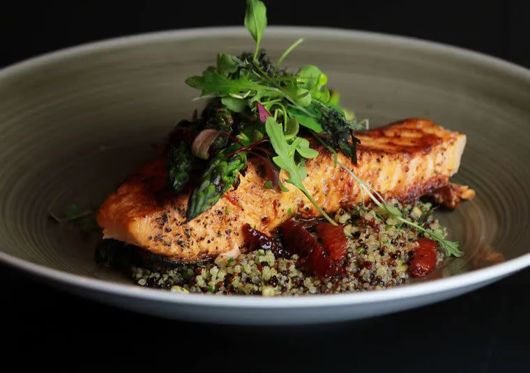 A grilled salmon fillet topped with microgreens sits on a bed of quinoa and asparagus, drizzled with a dark sauce, served on a greenish-gray plate against a dark background.