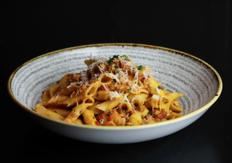A bowl of pasta topped with tomato-based sauce, grated cheese, and fresh herbs is served in a rustic white, shallow bowl against a black background.
