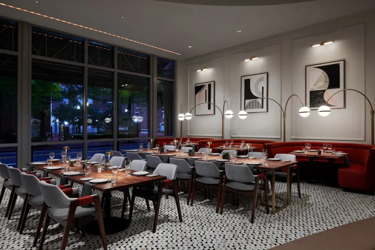 A modern restaurant dining area with large windows, red booth seating, gray chairs, set wooden tables, geometric tile floor, and abstract black-and-white artwork on the walls. Round pendant lights hang above the tables.