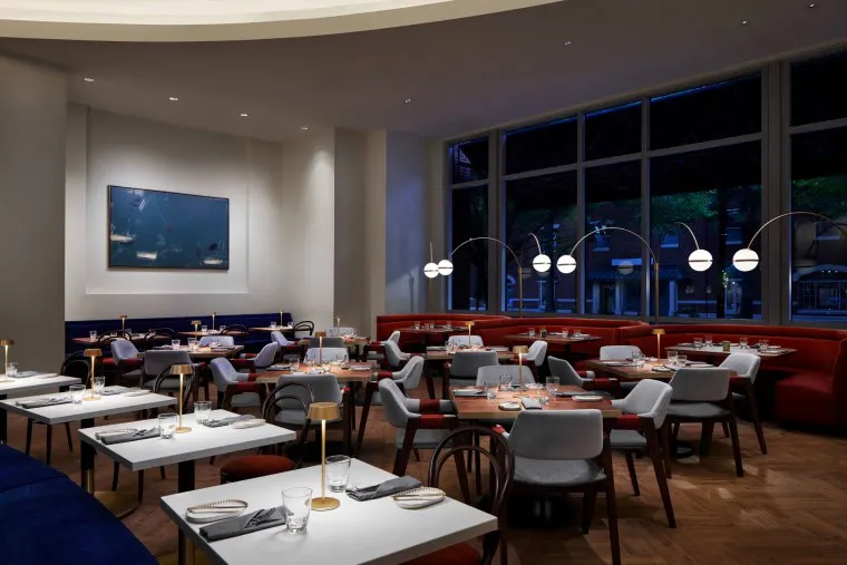 Modern restaurant interior with neatly set tables, cushioned chairs, red and blue banquette seating, stylish lamps, large windows, and contemporary wall art, creating a cozy and elegant dining atmosphere.