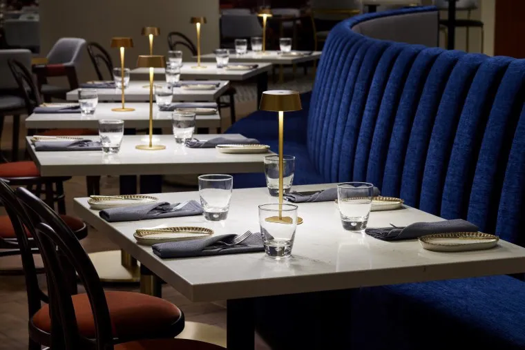 Elegant restaurant tables set with plates, glasses, and folded napkins. Small gold table lamps provide warm lighting. A plush blue banquette lines one side, with brown chairs on the other. The atmosphere is modern and sophisticated.
