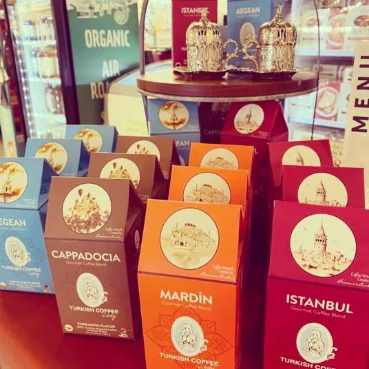 A display of colorful boxes of Turkish coffee blends labeled Cappadocia, Mardin, Istanbul, and Aegean. Decorative metal teapots sit on a tray above the boxes. A refrigerator and menu are visible in the background.