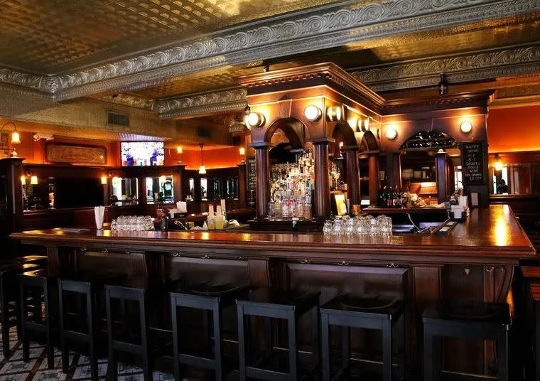 A dimly lit bar with a polished wooden counter, high wooden stools, shelves of liquor bottles, and glassware. Ornate molding and warm wall lights create a classic, inviting pub atmosphere.