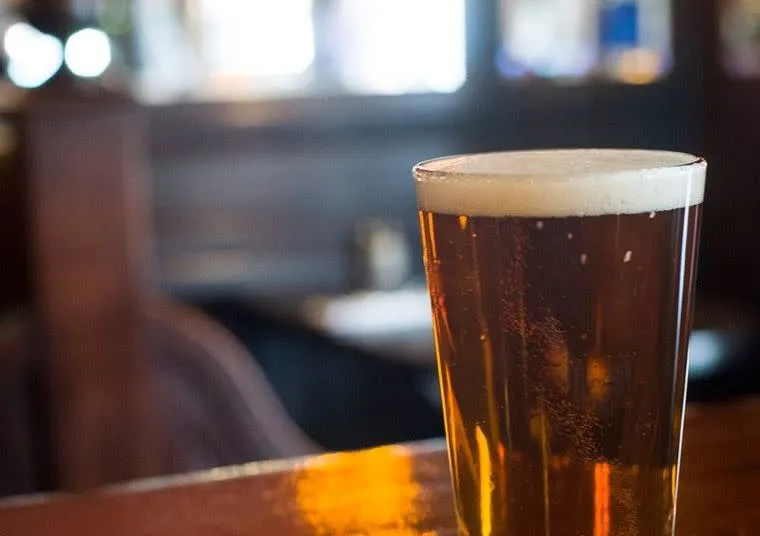 A pint glass filled with amber-colored beer topped with a foamy head sits on a wooden bar counter in a dimly lit pub.