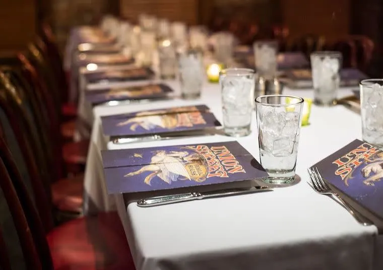 A long dining table set for a meal with glasses of ice water, silverware, and colorful menus on white tablecloths. The chairs are arranged neatly along both sides of the table.