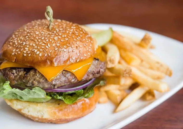 A cheeseburger with lettuce, red onion, and melted cheddar on a sesame seed bun sits on a white plate next to a side of golden French fries and a pickle spear.