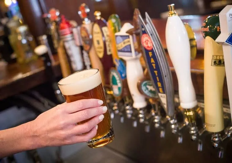 A hand holds a pint of draft beer in front of a row of colorful beer taps at a bar. The glass is filled with amber beer and a frothy head. The background shows various labels on the taps.
