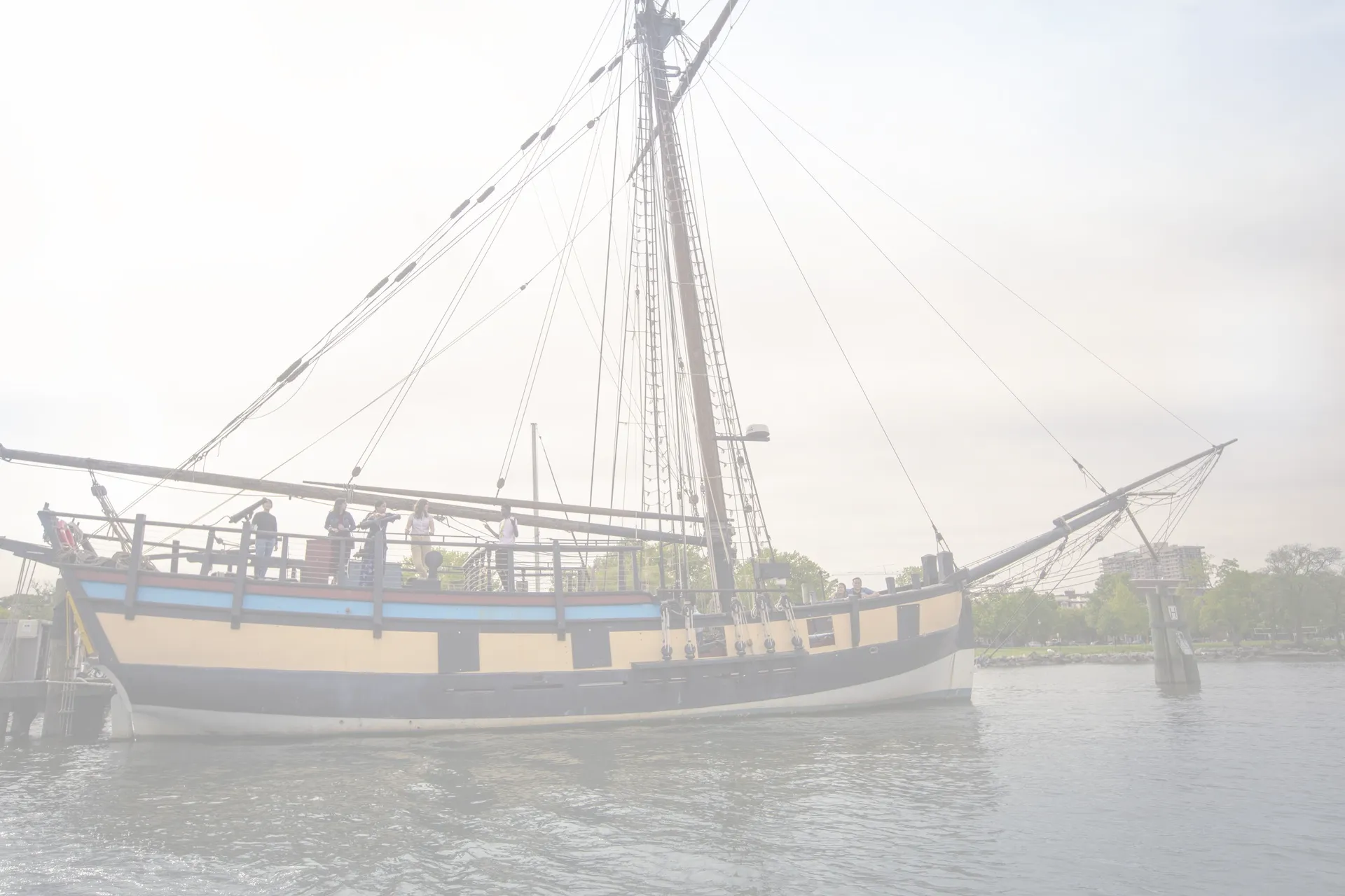 A large wooden sailing ship with tall masts and rigging is docked by the water, with several people standing on its deck. Trees and a pier are visible in the background.