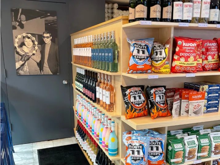 A store display with shelves of snacks, chips, and bottled drinks. On the left wall, theres a black and white photo of two people smiling and wearing sunglasses. Bottles and chips are arranged neatly on the shelves.