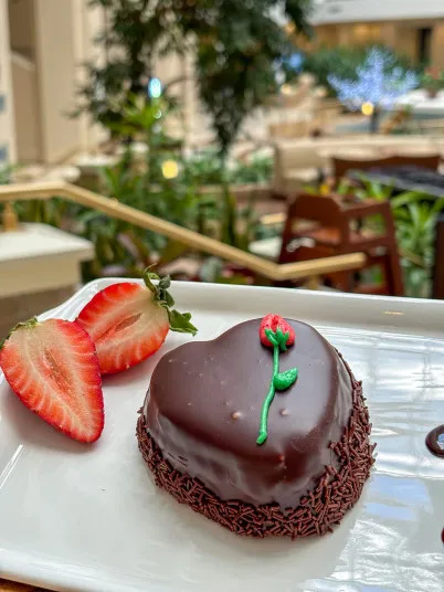 A heart-shaped chocolate dessert decorated with a small red rose design sits on a white plate next to two sliced strawberries. The background shows a blurred, green, indoor garden setting.