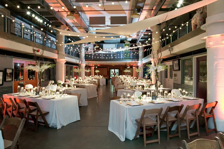 A modern, indoor wedding reception with long tables covered in white tablecloths, elegant place settings, tall floral centerpieces, wooden chairs, and string lights hanging from the ceiling.