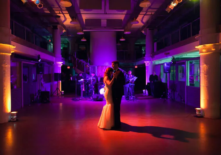 A bride and groom share a romantic first dance on an empty, dimly lit dance floor, surrounded by purple and orange uplighting, with a live band playing in the background.