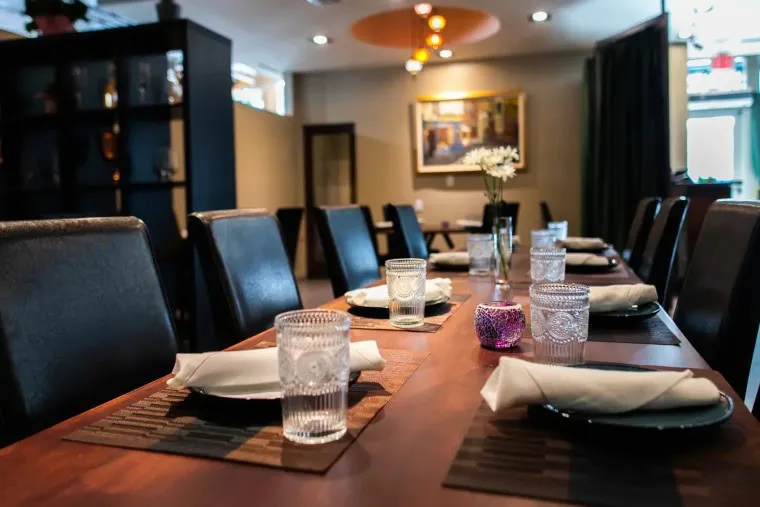 A long restaurant table is set with glasses, white napkins on plates, and small vases with flowers. The dining area has warm lighting, dark chairs, and modern decor, creating a welcoming atmosphere.