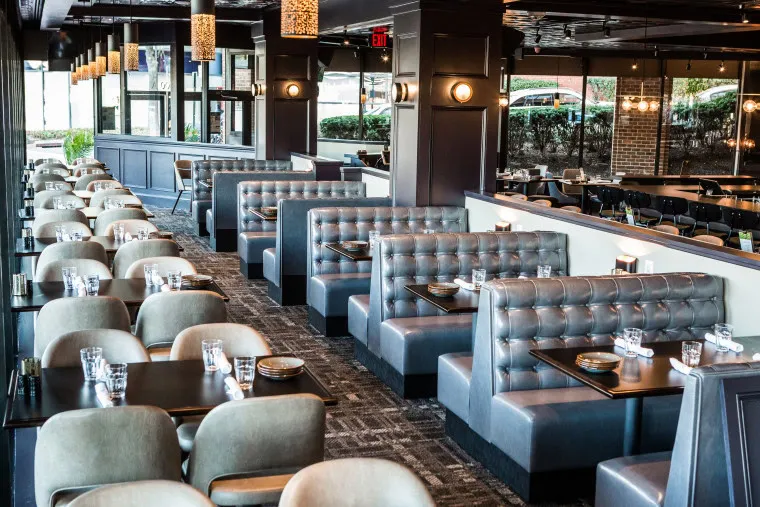 Modern restaurant interior with empty gray upholstered booths and tables, neatly set with plates, glasses, and silverware. Large windows line one side, letting in natural light, and pendant lights hang from the ceiling.