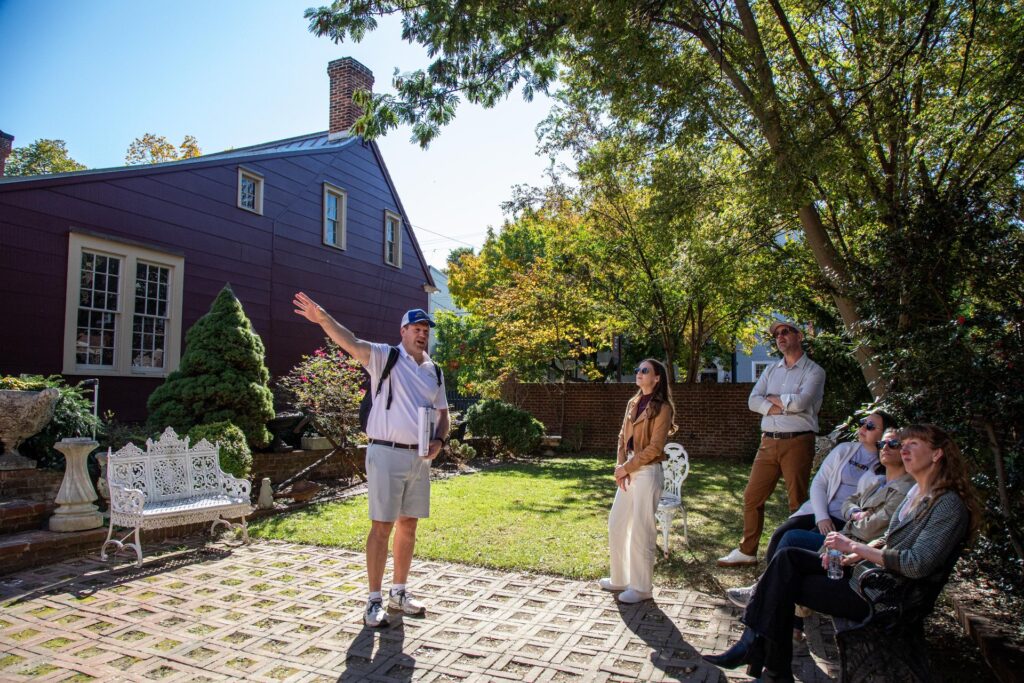 A man giving a guided outdoor tour gestures while a group listens attentively in a sunny garden with benches, trees, and a red house—one of the best things to do this summer for those seeking relaxation and discovery.