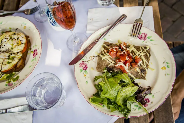 A plate with a buckwheat crepe topped with diced tomatoes and cheese, mixed greens, a glass of iced water, a glass of red drink, and another plate with toast and cheese, all on a wooden table with a white cloth.