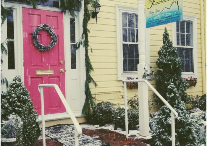 A bright pink door with a wreath is decorated with greenery and snowy plants outside. Yellow siding, a sign reading Day Spa, and white railings create a welcoming winter scene. Snow is gently falling.