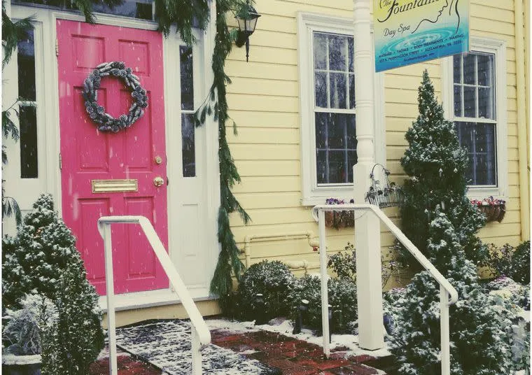 A bright pink door with a wreath is decorated with greenery and snowy plants outside. Yellow siding, a sign reading Day Spa, and white railings create a welcoming winter scene. Snow is gently falling.