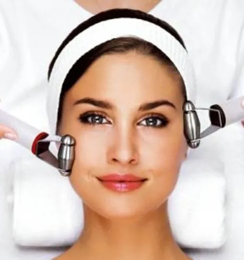 A woman with a white headband is receiving a facial treatment using two metal massage devices on her cheeks. She has clear skin and is lying on a spa bed with a towel behind her head.