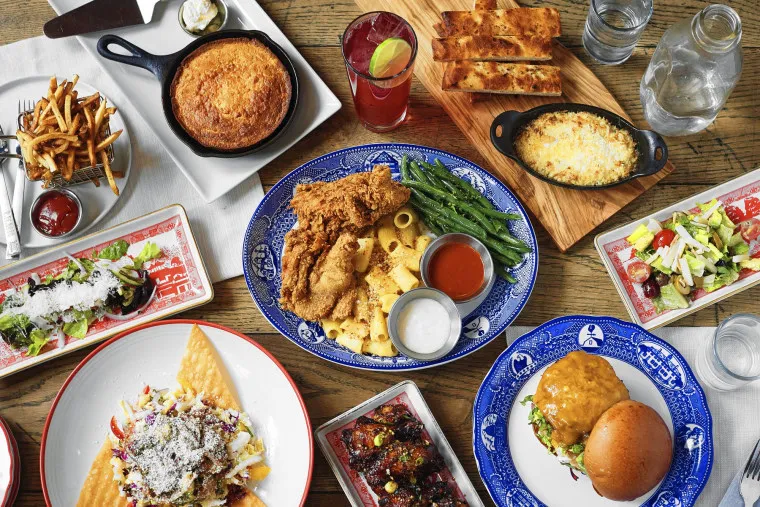 A table filled with assorted dishes including fried chicken with green beans and pasta, burgers, cornbread in a skillet, fries, flatbread, salad, cheesy dip, and drinks, all set on wooden surface.