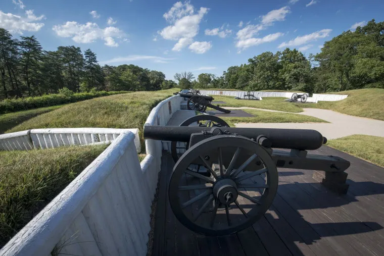 Several historic cannons are lined up behind white wooden barriers on an earthwork fort under a blue sky with scattered clouds and surrounding green trees.