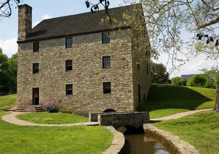 A historic stone building with a dark roof stands on green grass. A narrow stream runs under a small stone bridge in front, with trees and a partly cloudy sky in the background.