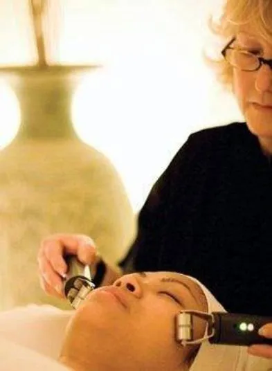 A person receives a facial treatment with two handheld devices operated by a professional in a softly lit spa setting.